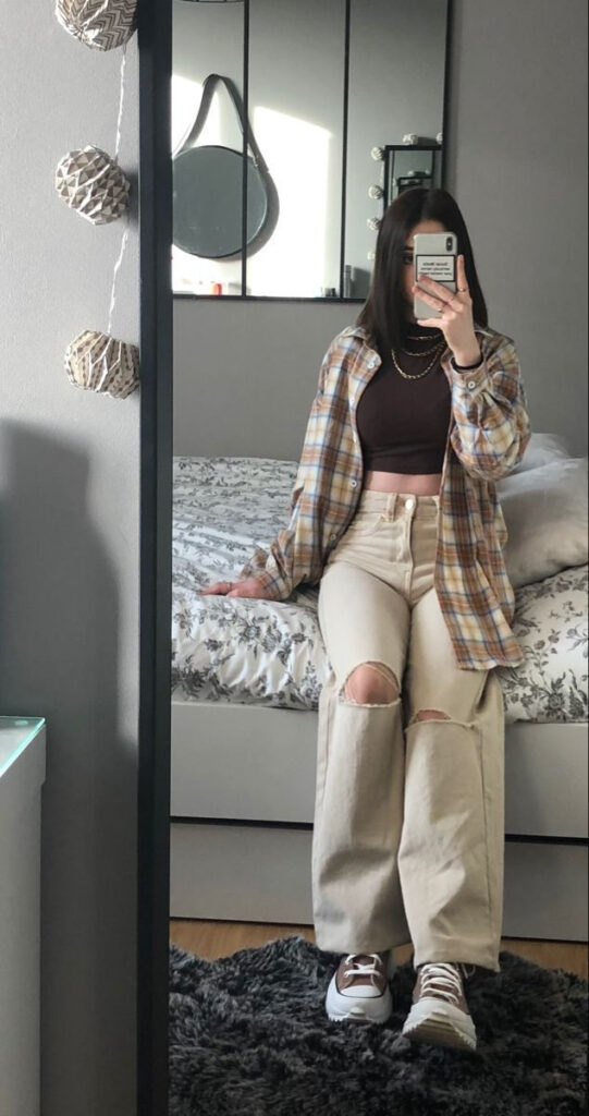 A girl takes a full-length mirror selfie while sitting on the edge of a bed. she is wearing distressed high-waisted beige wide-leg jeans with ripped knees, a brown crop top, and an open plaid flannel shirt. They are wearing chunky brown sneakers with thick white soles and a layered gold chain necklace.