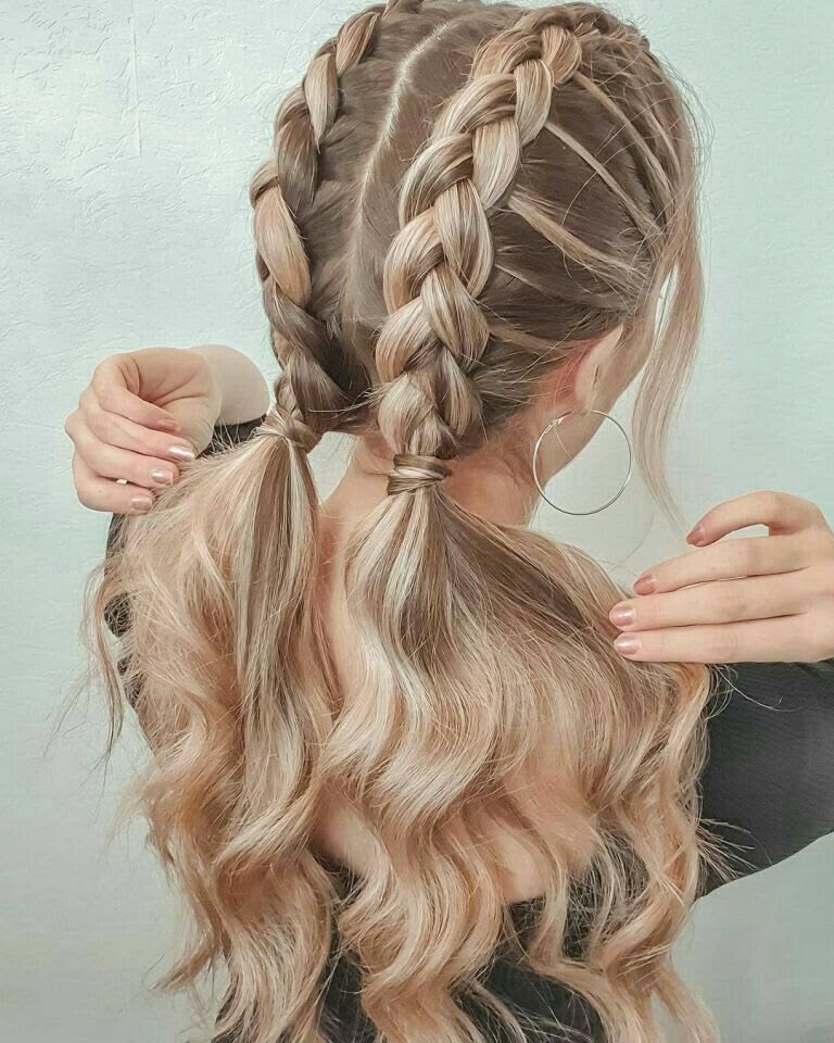 A back view of a woman with blonde highlighted hair featuring two French braids that turn into low pigtails with loose, wavy ends.