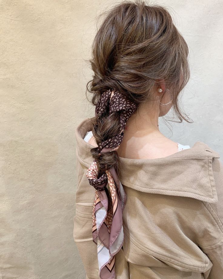 Side and back view of a woman with messy, textured brown hair showcasing how to style a bandana or scarf. A brown paisley print silk scarf is woven directly into a loose low braid, cascading down her back over a beige jacket.