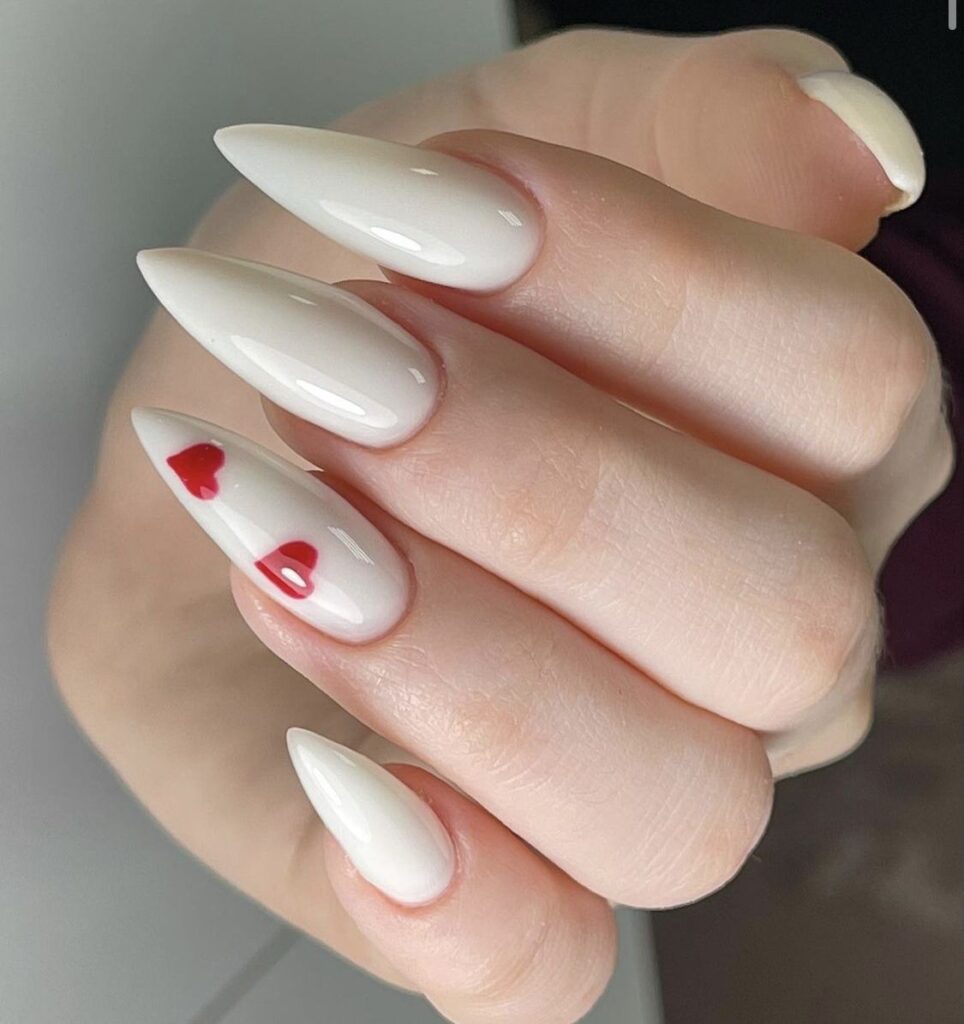 Long, sharp stiletto-shaped nails painted in a high-gloss milky white color, featuring a single accent nail with two small glossy red hearts painted vertically in the center.