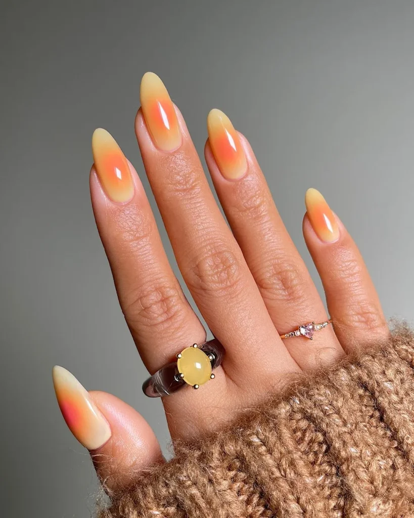 Long almond-shaped nails with a high-gloss finish, featuring a creamy pastel yellow base coat decorated with a vibrant coral-orange airbrushed aura design in the center of each nail.