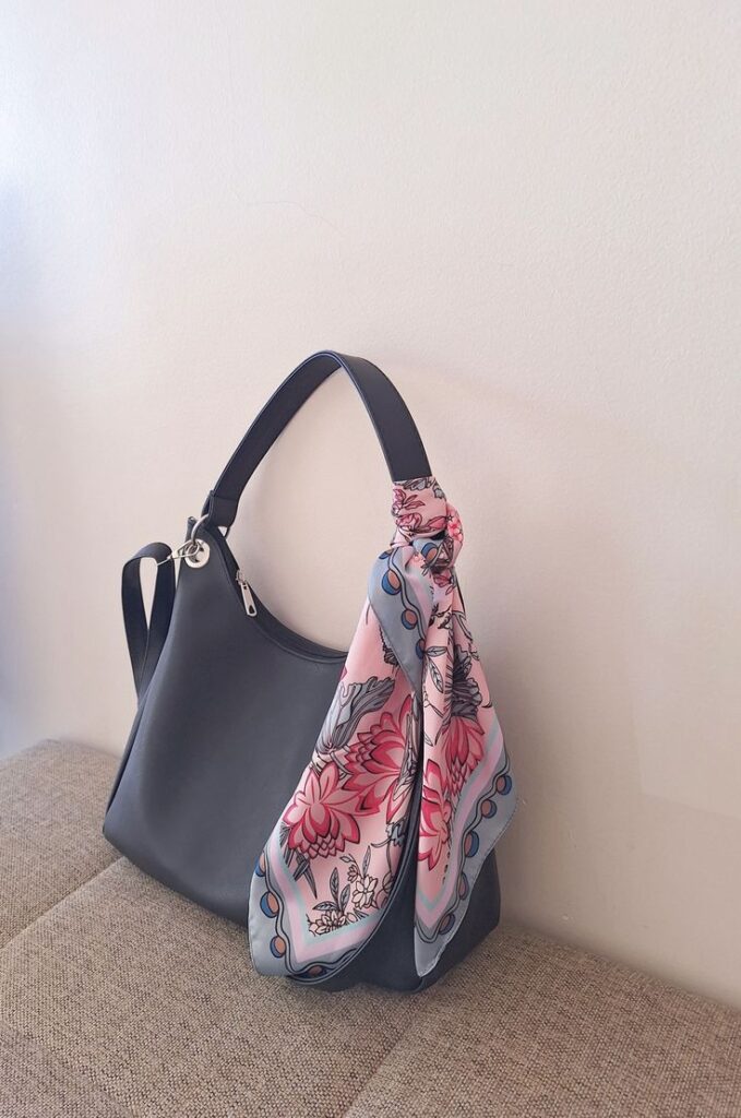 A gray leather shoulder bag showing how to style a bandana or scarf. A pink floral silk scarf is tied around the base of the bag handle.