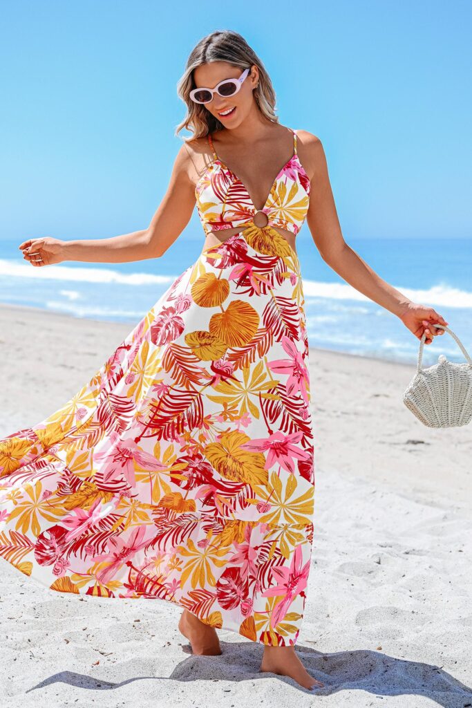 A smiling woman standing on a beach wearing a tropical cutout maxi dress, white sunglasses, and holding a small white bag for beach vacation outfits.