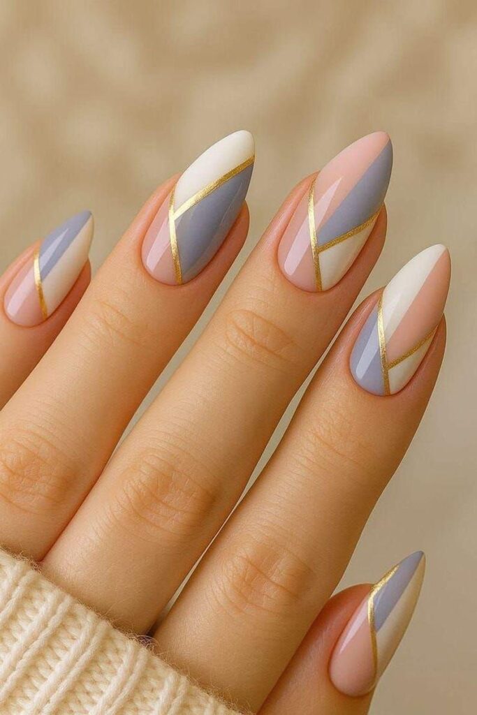 Almond-shaped manicured nails with a clear base, decorated with geometric abstract color blocks in pastel pink, lavender gray, and white, all separated by thin, metallic gold lines.