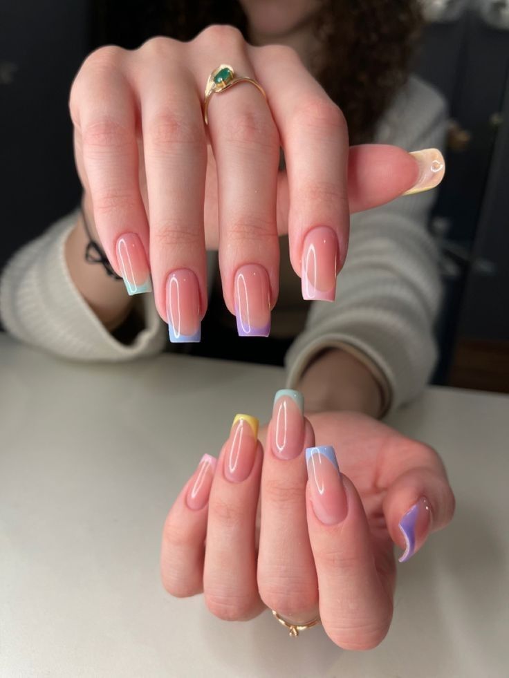 A close-up of a girl's hands showing long square nails with a nude base and pastel rainbow French tips, featuring soft pink, blue, purple, mint green, and yellow shades.