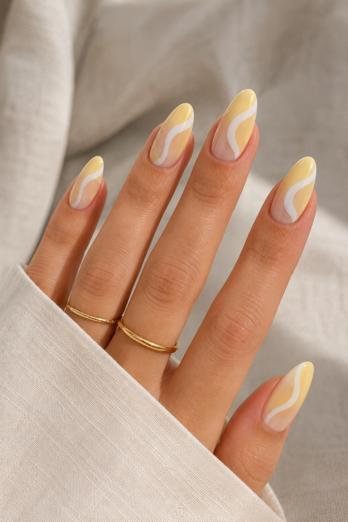 A fresh, bright close-up showing a chic abstract manicure on almond nails. Soft yellow and stark white ribbon-like waves twist across a clear base coat. The hand is accessorized with two thin gold bands against a neutral fabric background.