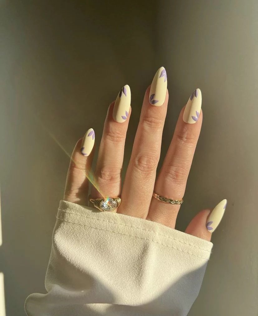 A high-quality lifestyle shot under warm, direct sunlight showing a hand with long, pointed almond nails. The manicure features a solid pastel butter yellow base with small, elegant purple leaf art painted on the sides. The hand wears a large crystal statement ring and a gold band, peeking from a cream cuff.