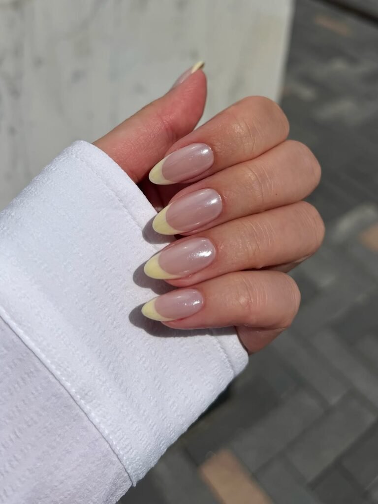 A close-up of a hand showcasing almond-shaped nails with a pastel butter yellow French tip manicure. The nails have a high-gloss, slightly iridescent glazed finish over a natural nude base. The person is wearing a crisp white button-down shirt sleeve.