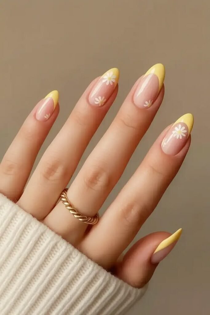 A high-quality close-up of almond-shaped nails with a smooth nude base and light butter yellow French tips. The design is beautifully decorated with small, hand-painted white daisy flowers, accessorized with a twisted gold ring and a white sweater cuff.