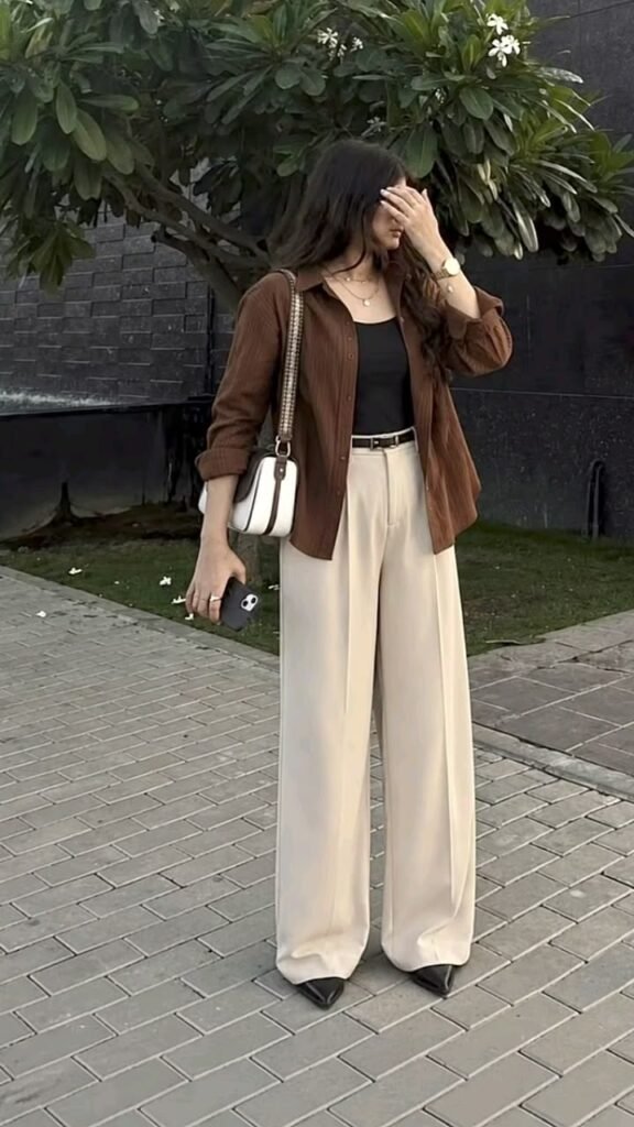 Woman wearing cream wide leg trousers with a black tank top, brown lightweight shirt, pointed-toe flats, and a shoulder bag for a business casual summer outfit.