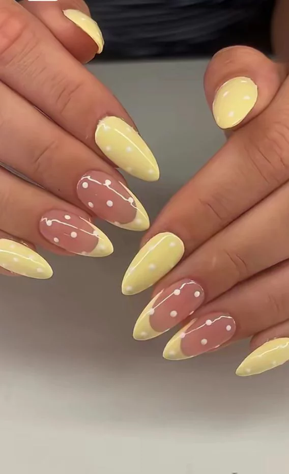 A close-up of almond-shaped nails with a light butter yellow and white polka dot design. Some nails are fully painted yellow, while others have yellow French tips on a natural base, all decorated with small white dots.