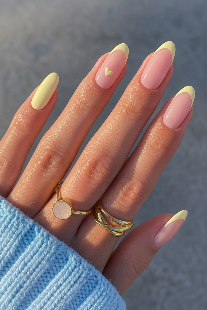 A high-quality close-up of almond-shaped nails with a light butter yellow French tip manicure. The design includes a sheer nude base and a tiny yellow heart painted on the middle finger, accessorized with gold rings and a blue knit sweater.