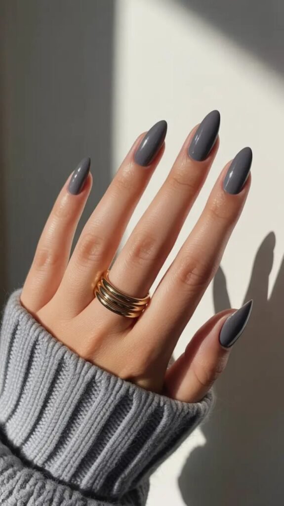 Glossy charcoal gray almond-shaped nails with a smooth gel finish, styled with a cozy gray knit sweater and chunky gold rings for a chic minimalist manicure.