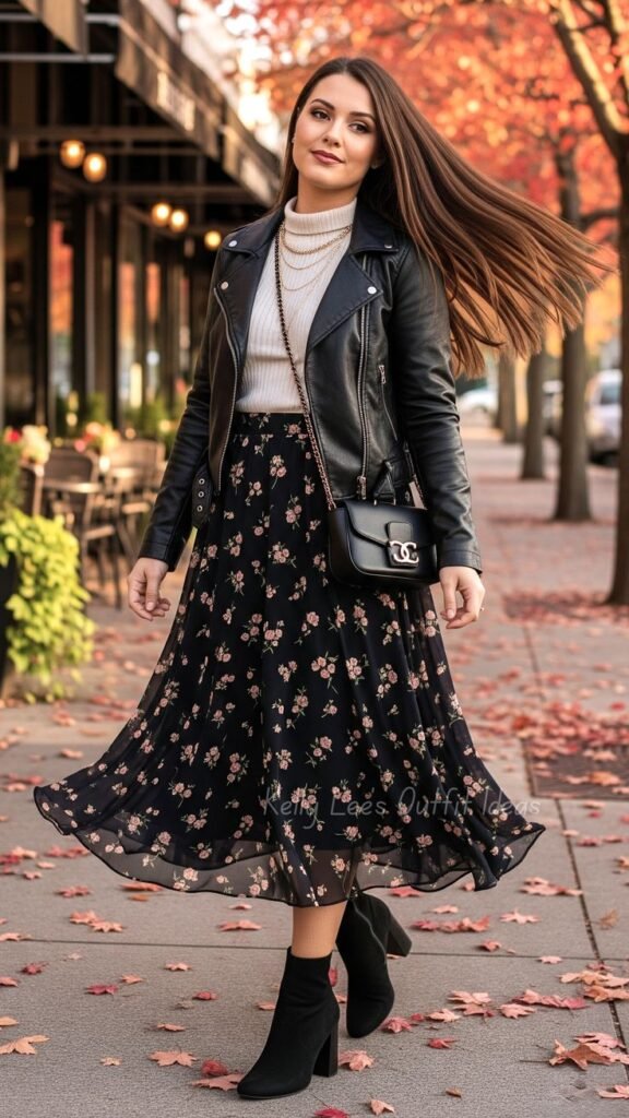 Woman walking down an autumn street wearing a black leather moto jacket, cream mock-neck sweater, black pink-floral midi skirt, and black heeled ankle boots.