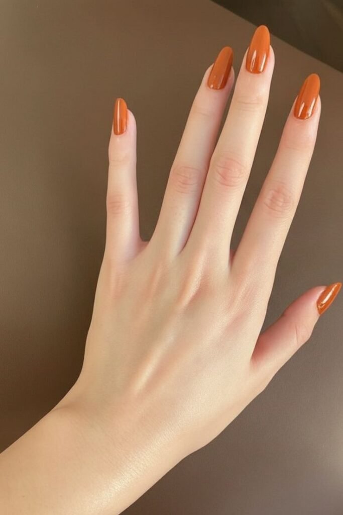 Glossy terracotta orange fall nails, minimalist pumpkin spice almond manicure, and simple solid-colored autumn nail design.