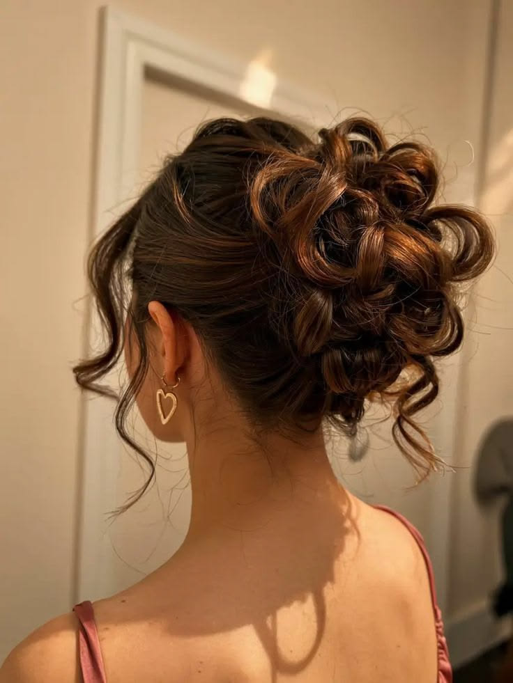 Back view of a young woman with brown hair styled in a romantic, messy curly updo. The hairstyle features pinned curls and loose, wavy tendrils framing her neck. She is wearing gold heart-shaped earrings.