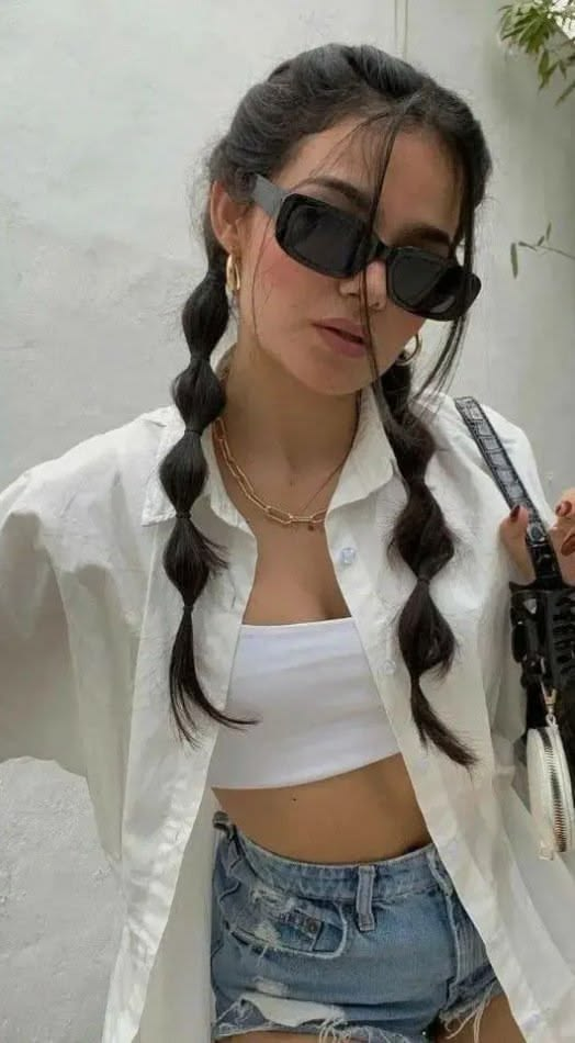 A young woman wearing sunglasses and a white open button-up shirt, showcasing a casual hairstyle with double bubble pigtails and loose strands framing her face.