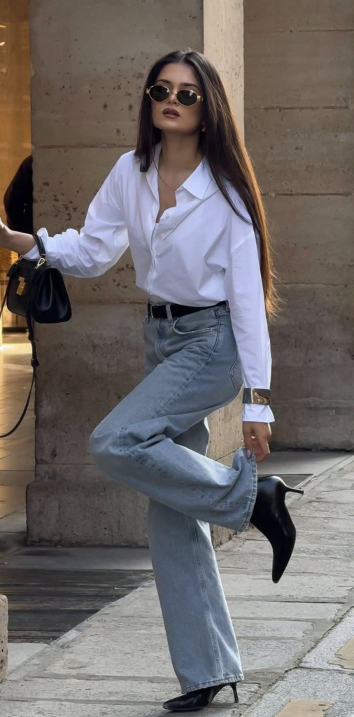 "Woman in a white button-down shirt, light-wash wide-leg jeans, and black pointed-toe heel boots holding a black leather handbag."