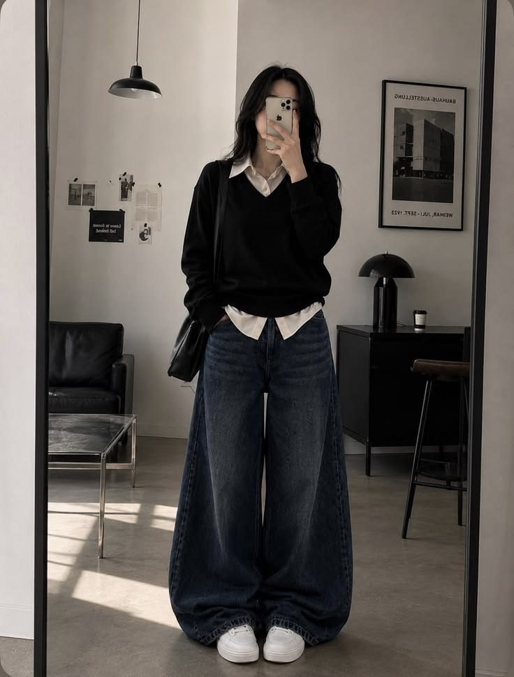 Woman taking a mirror selfie wearing a layered black V-neck sweater over a cream shirt, paired with very wide-leg dark jeans and white sneakers