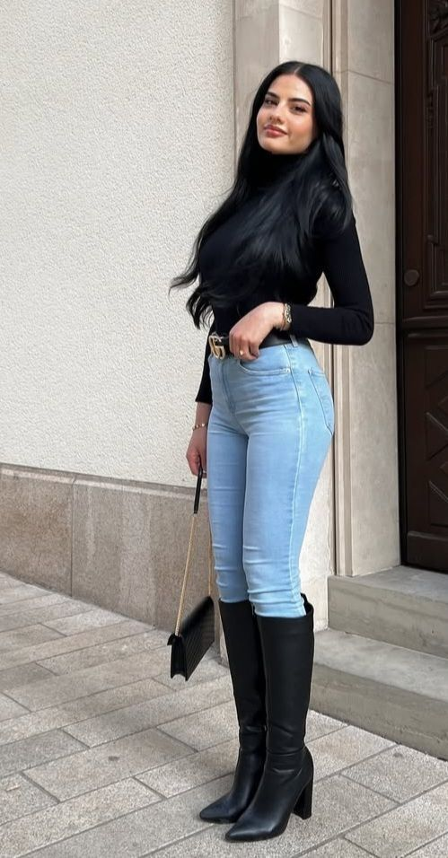 A woman posing outdoors wearing a black turtleneck sweater, light blue skinny jeans, and tall black leather heel boots perfectly giving inspiration for jeans outfit ideas.