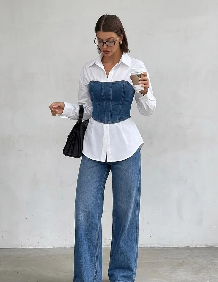 A woman wearing a white collared shirt layered under a blue denim corset, paired with loose blue jeans and holding a black bag.
