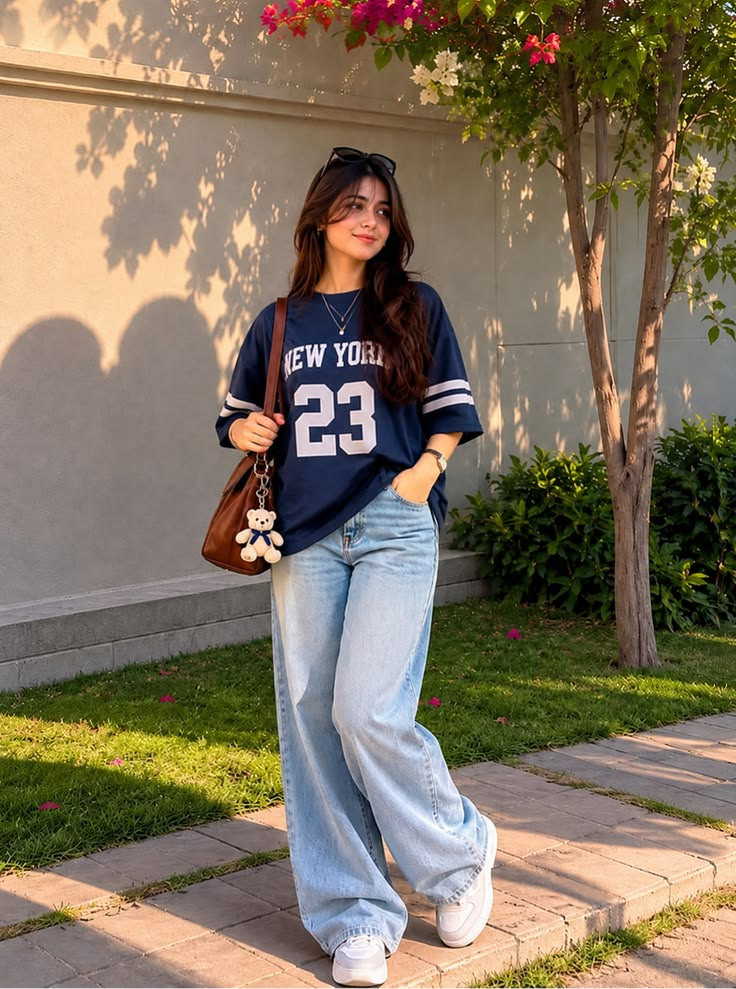 Chic casual street style look with a navy graphic tee, baggy light blue jeans, white shoes, and a brown bag.