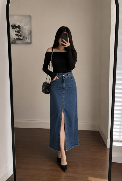 A woman taking a mirror selfie wearing a fitted black off-the-shoulder top, a high-waisted denim maxi skirt with a center slit, pointed-toe black heels, and a black chain shoulder bag.