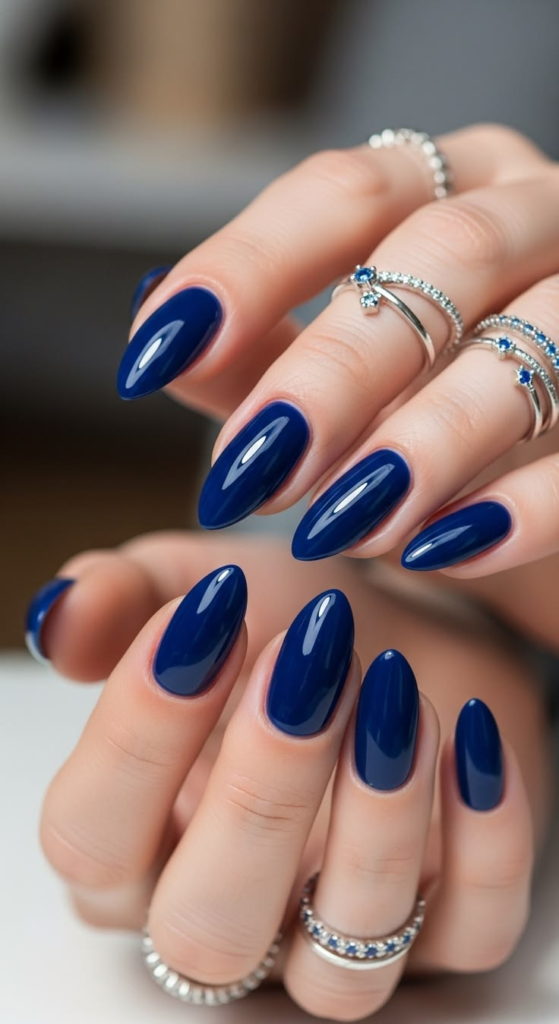 Glossy navy blue nails, solid royal dark blue cream manicure on medium almond nails, and elegant minimalist midnight blue nail design with silver ring jewelry.
