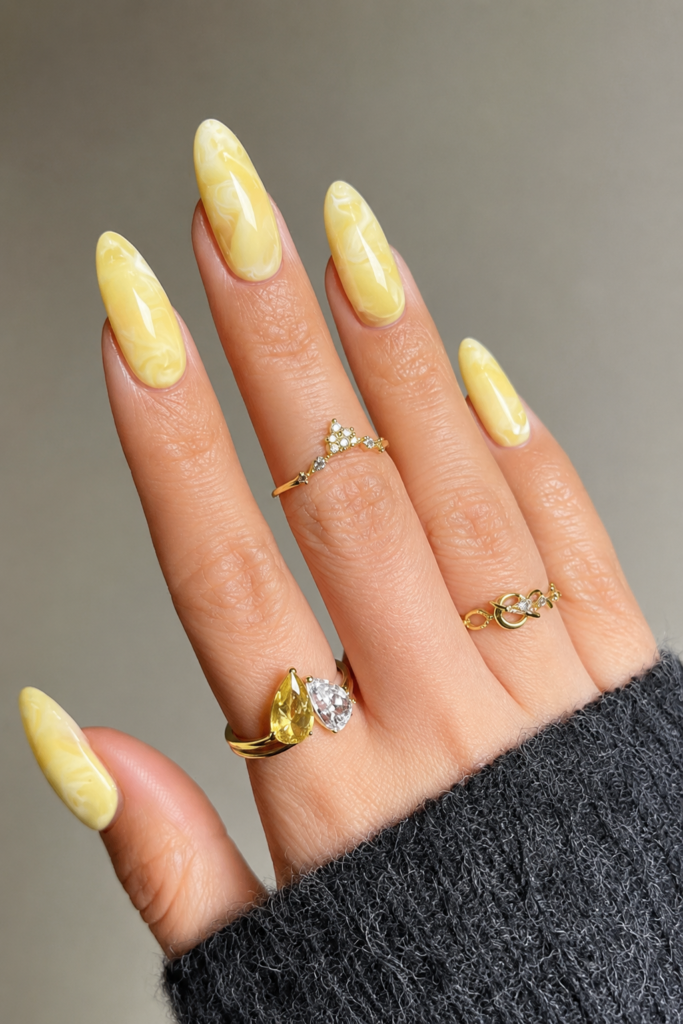 A high-quality close-up of long almond-shaped nails with a light butter yellow and white marble texture design. The hand is decorated with delicate gold rings, including one with a yellow gemstone, and wears a dark gray knit sweater.
