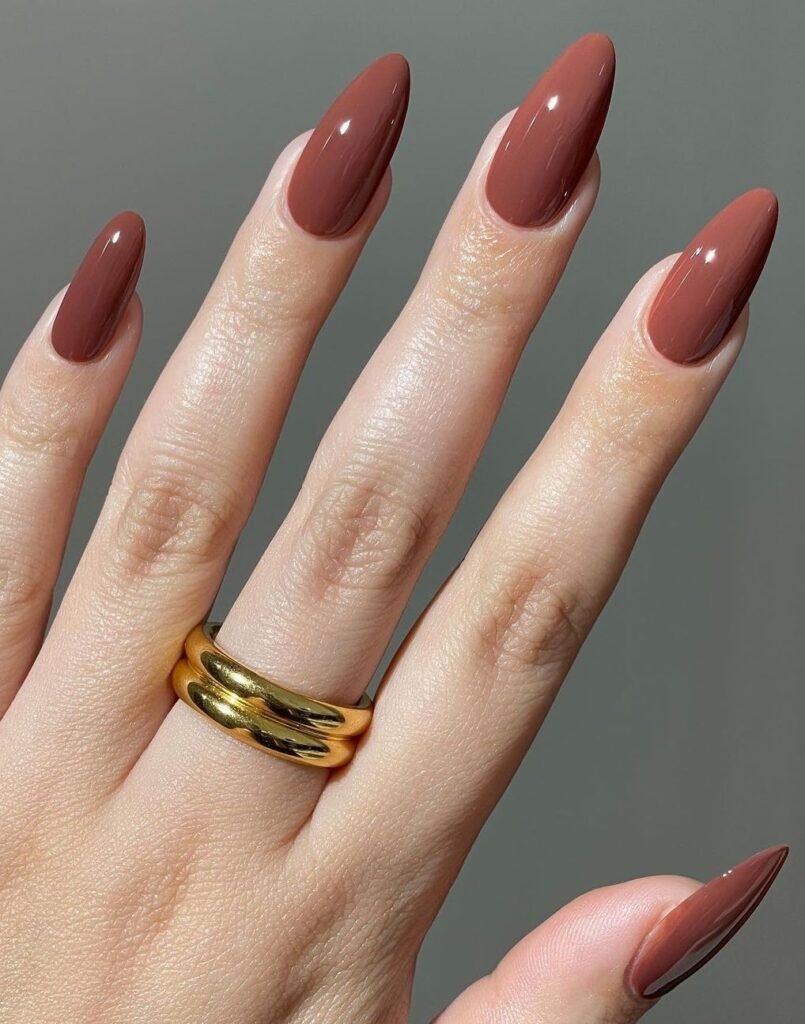 Glossy mocha brown almond-shaped nails with a minimalist solid color manicure and gold statement ring.