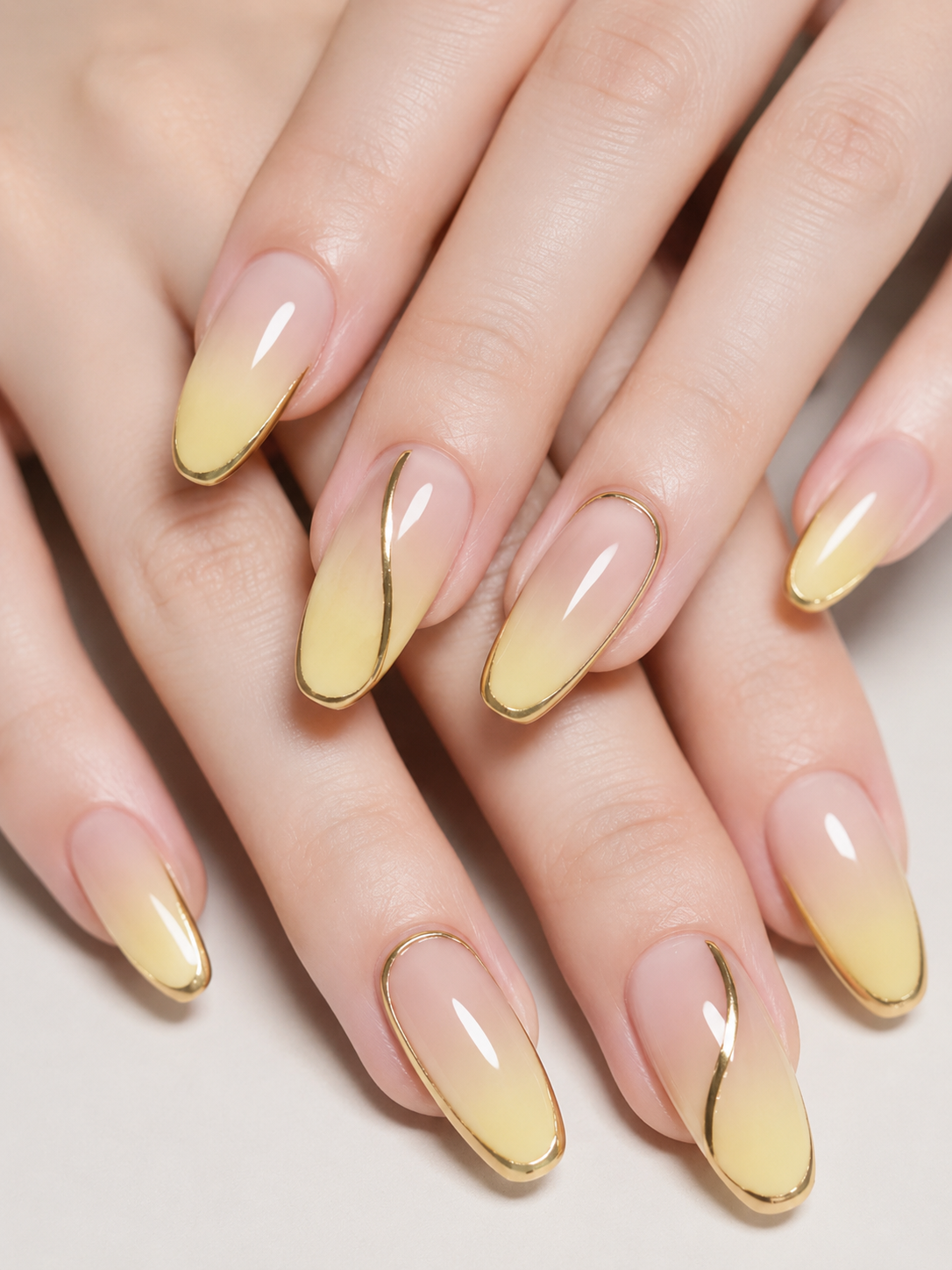 A professional close-up of a hand with long, elegant almond and oval-shaped nails. The manicure features a smooth nude-to-butter-yellow ombre gradient, intricately detailed with thin, shiny metallic gold chrome outlines and delicate wavy gold accents.