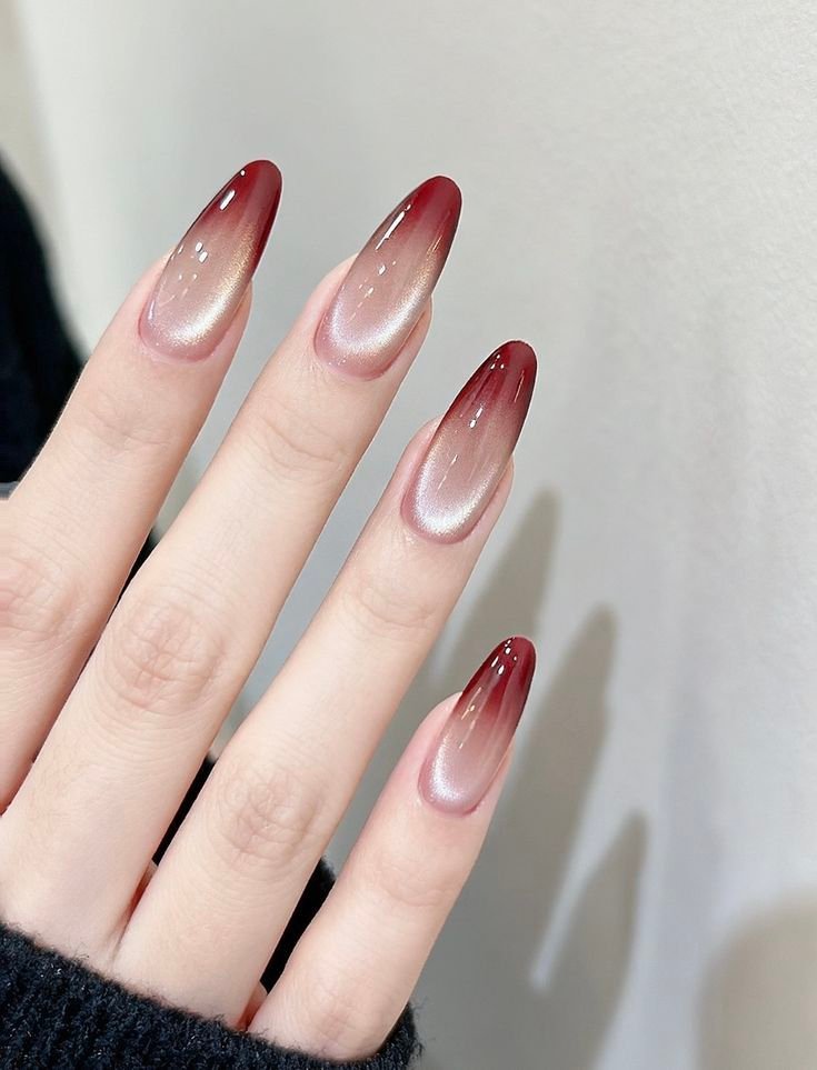 Long, elegant oval nails featuring a sheer nude base blending into deep red ombre tips, topped with a glowing champagne velvet magnetic cat eye nail designs effect.