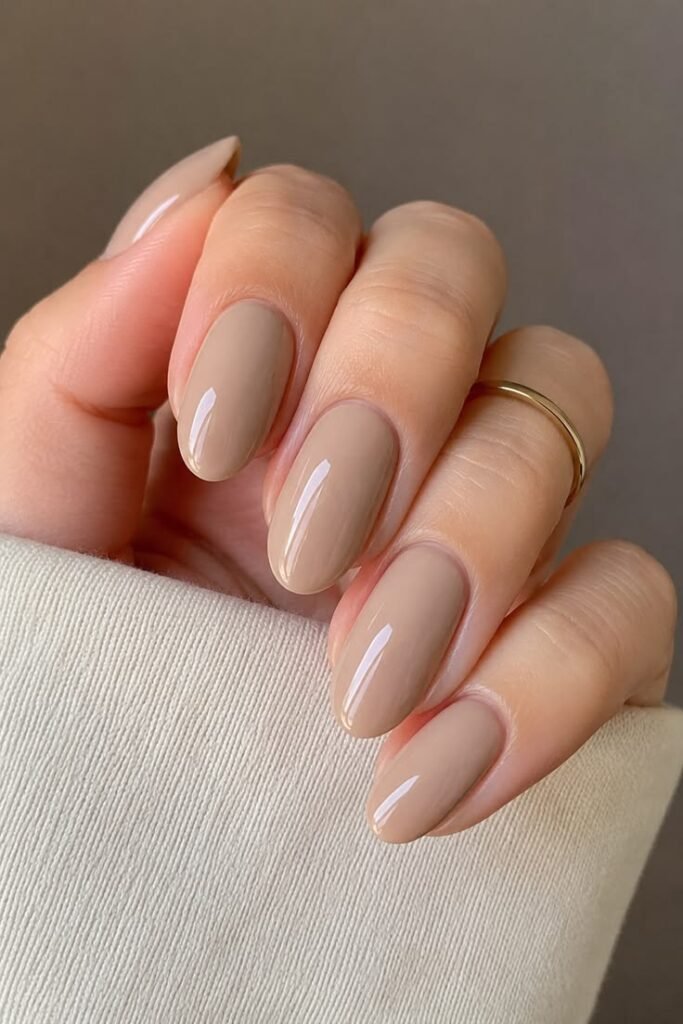 Glossy nude beige almond-shaped nails with a minimalist solid color manicure and gold ring.