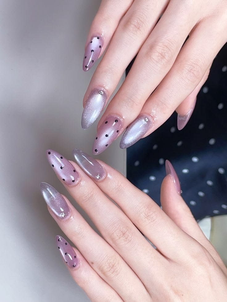 Long, pointed stiletto nails featuring alternating designs of sheer lavender with black polka dots and shimmering silver-purple velvet magnetic cat eye nail designs accented with small rhinestones.
