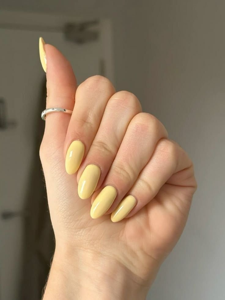 Close-up of glossy solid butter yellow almond-shaped nails with a smooth pastel finish, photographed on a hand wearing a simple silver ring against a soft neutral background.