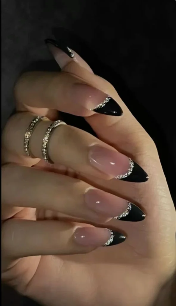 A close-up beauty shot of luxurious nail designs featuring almond-shaped nude nails with glossy black French tips separated by a sparkling silver glitter line.