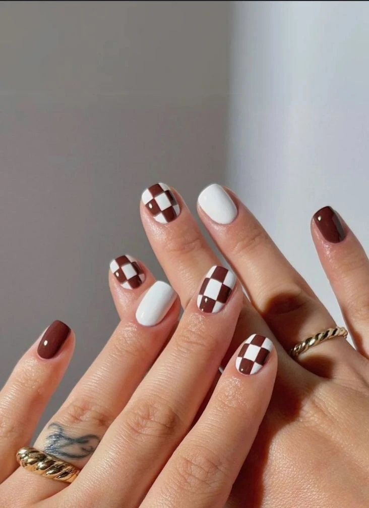 A close-up beauty shot of cute and playful short nail ideas featuring short squared-square nails with solid brown, solid white, and brown-and-white checkerboard accent patterns.