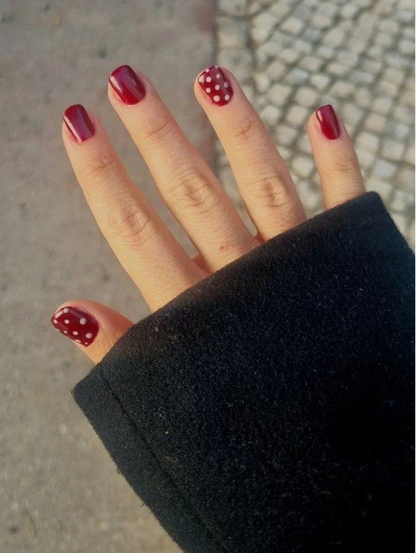 A close-up lifestyle shot of short nail ideas featuring a glossy deep burgundy manicure on short squoval nails with white polka dot accent details, paired with a black coat.