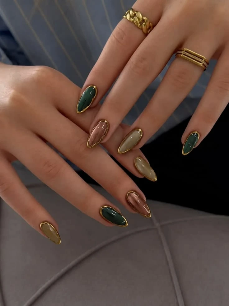 A close-up view of an elegant luxury manicure featuring almond-shaped nails in rich emerald, olive, and metallic taupe tones with raised gold chrome frames, creating a sophisticated jewelry-inspired nail art design.