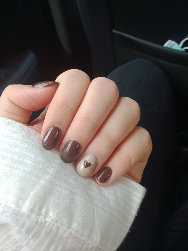 A close-up lifestyle shot of short nail ideas featuring an espresso brown manicure on short squoval nails, a creamy beige accent nail with a small brown heart design, and a white textured sleeve.