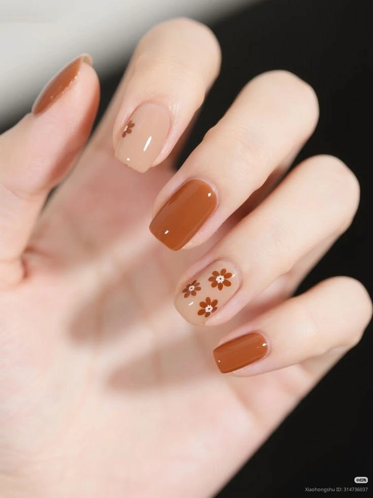 A close-up beauty shot of short nail ideas featuring a glossy caramel brown manicure on short squoval nails with creamy nude accent nails decorated with brown daisy flower art.