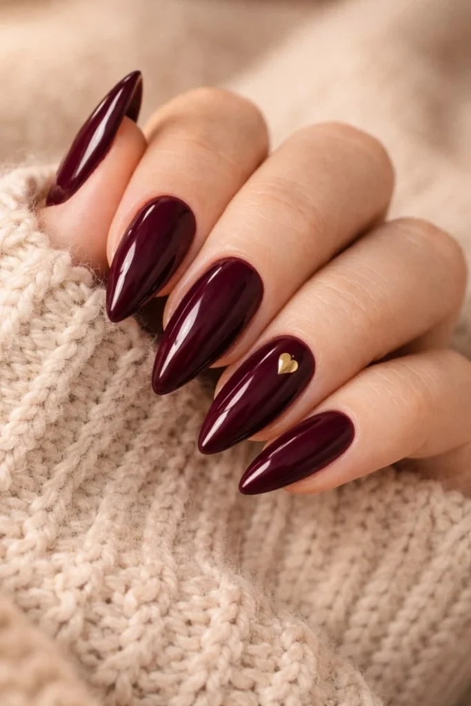 A close-up luxury beauty shot of trendy and luxurious nail designs featuring long almond nails in rich glossy burgundy with a tiny gold heart accent, resting against a cozy cream knit sweater.