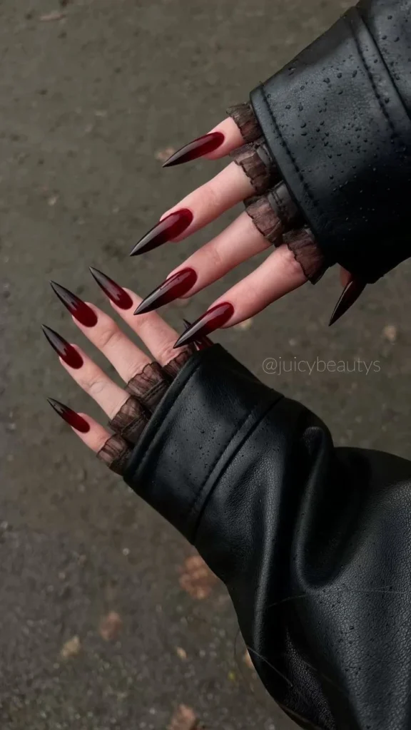 A close-up beauty shot of fierce stiletto-shaped nails featuring a deep red to black ombre design with a glossy finish, perfect for a bold gothic-inspired luxury manicure.