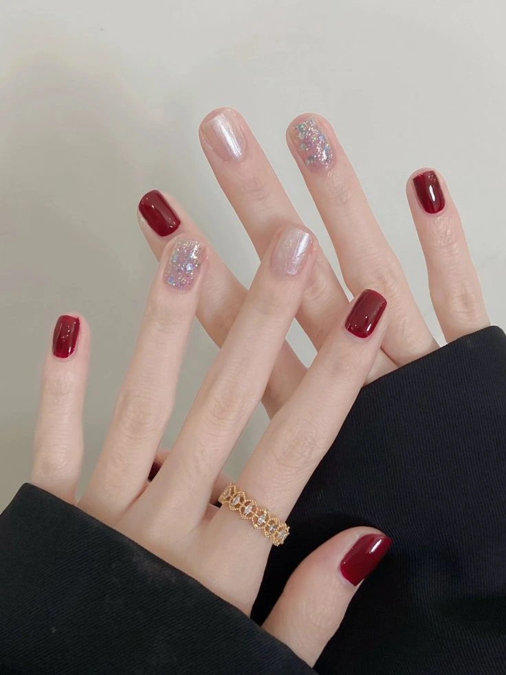 A close-up beauty shot of trendy and luxurious short nail ideas featuring short squared-oval nails with glossy deep red polish, pearlescent chrome, and chunky multi-colored glitter accents, styled with a gold band ring.