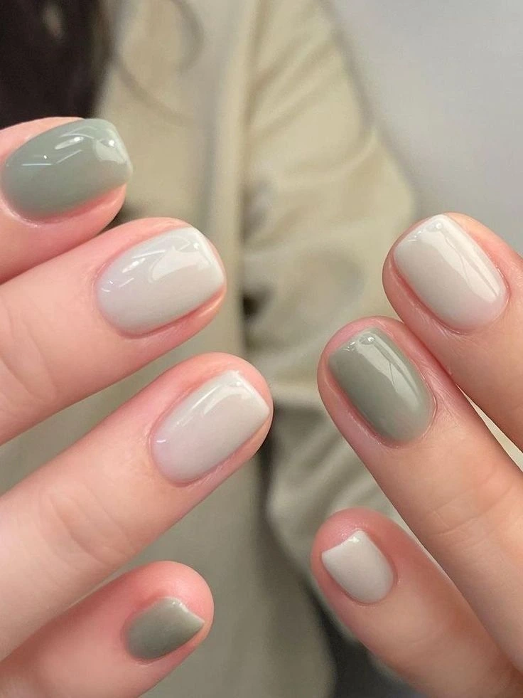 An aesthetic macro shot of short square nails featuring a muted skittle manicure design. The fingers showcase alternating dusty sage green and warm stone grey gloss polishes with a smooth gel texture, styled elegantly against a cream-colored soft fabric coat background for a sophisticated autumn fashion vibe.