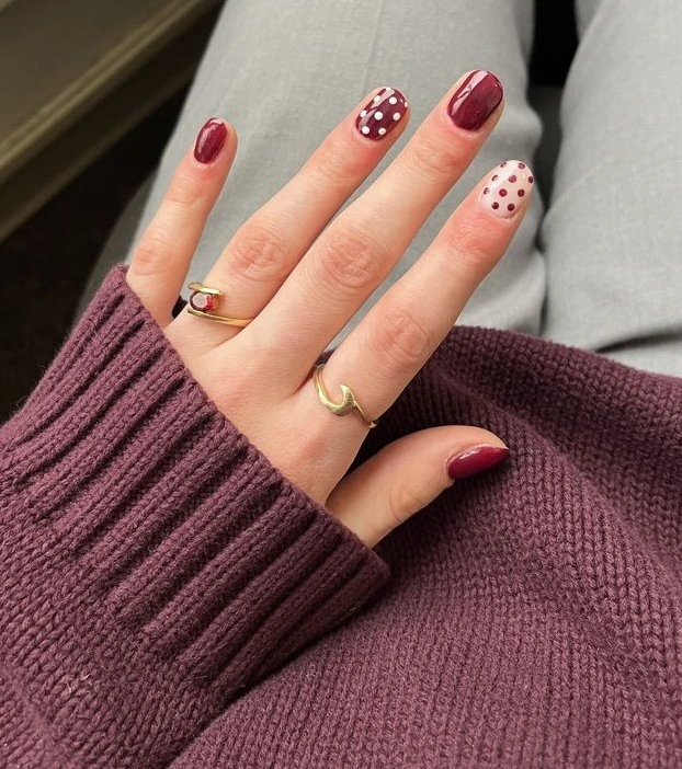 A close-up lifestyle shot of a retro burgundy polka dot manicure on short squoval nails, paired with a cozy plum sweater and gold rings.