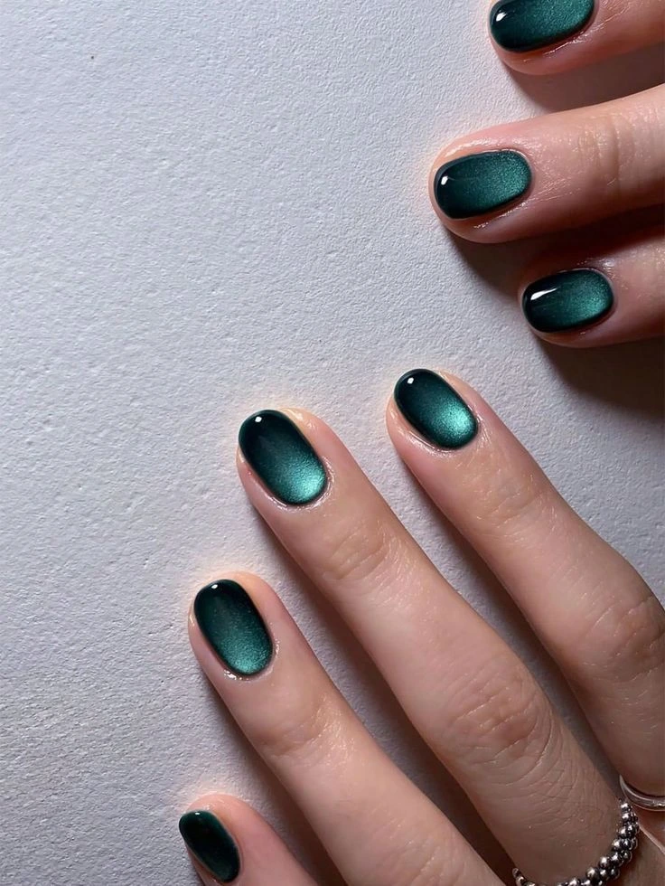A close-up beauty shot of trendy and luxurious short nail ideas featuring short oval nails with deep emerald green cat-eye magnetic gel polish creating a luminous velvet finish.