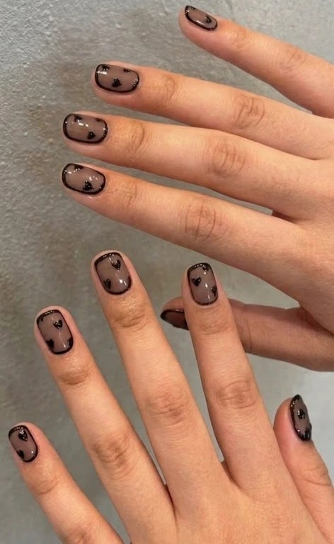 A close-up beauty shot of trendy short nail ideas featuring short square nails with a semi-sheer nude base, thin black outer frames, and tiny black heart designs.