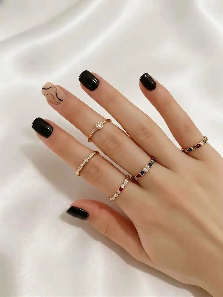 A luxury beauty close-up of a minimalist abstract manicure on short squoval nails. Four fingers feature a flawless, high-gloss opaque deep black polish, while the index accent nail showcases a chic nude-beige base with fine-line black organic swirls, adorned with multiple delicate gold gemstone rings over a premium white satin backdrop.