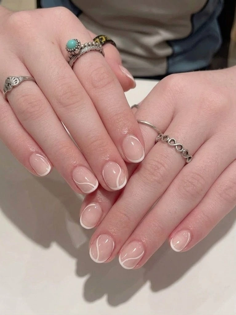 A detailed close-up shot of a minimalist abstract swirl manicure on perfectly short round nails. The hands display a sheer translucent nude pink base coat decorated with thin, fine hand-painted white wavy lines and micro French tips, accessorized elegantly with silver bohemian rings against a clean neutral background for a premium beauty blogger look.